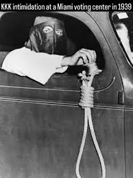 a hooded Klansman sitting casually beside a car near a Miami voting center